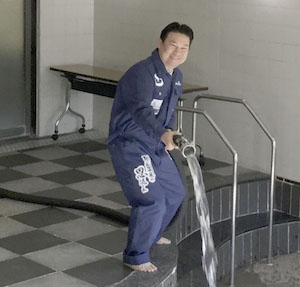 つなぎの作業着を着てホースを持ち、温泉の浴槽に湯をそそぎ入れる長野市長の写真