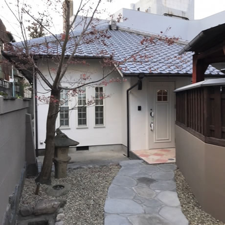 外観写真。日本的な石どうろうのある玉砂利をしいた庭に石畳の小道があり、瓦屋根に白い壁を持つ和洋折衷な見た目の家へ続いている。