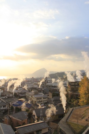 日が落ちかけた別府市街を小高いところから見た風景写真。湯けむりがそこかしこから立ち上っており、遠くには高崎山と別府湾が見えます。