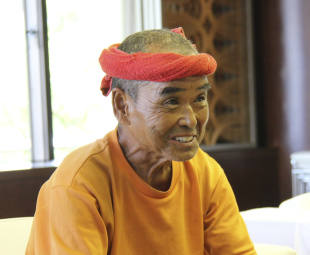 オレンジ色のTシャツと赤いタオルの針巻きをした、笑顔の尾畠春夫さんの写真